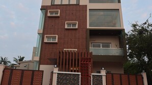A modern multi-story building with a mix of glass and wooden facade, a gated entrance, and two motorcycles parked in front. - MM Beach House (Tirupporur)