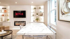 Un salon moderne avec une grande table en marbre blanc, une cheminée, un téléviseur affichant le logo « NETFLIX » et une bibliothèque agrémentée d’objets décoratifs.