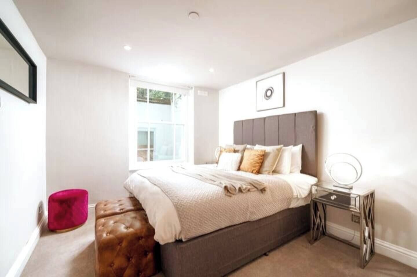 Un dormitorio con una cama grande, una mesita de noche con espejo, una ventana y una silla rosa.