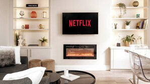 Un salon moderne doté d’une cheminée, d’un téléviseur fixé au mur qui diffuse du contenu « NETFLIX », et d’une bibliothèque contenant divers objets décoratifs.