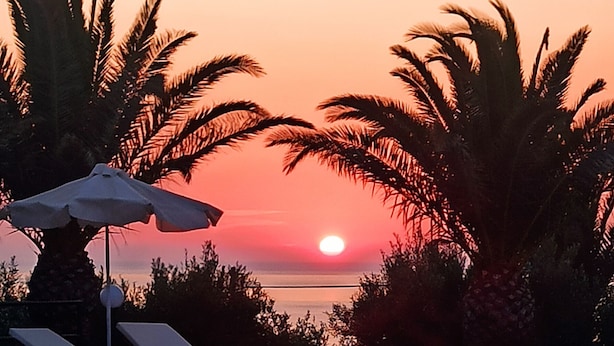 A swimming pool with lounge chairs and umbrellas, palm trees, and a sunset in the background. - Vida Balance Garden (Mithimna)