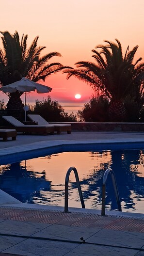 A swimming pool with lounge chairs and umbrellas, palm trees, and a sunset in the background. - Vida Balance Garden (Mithimna)