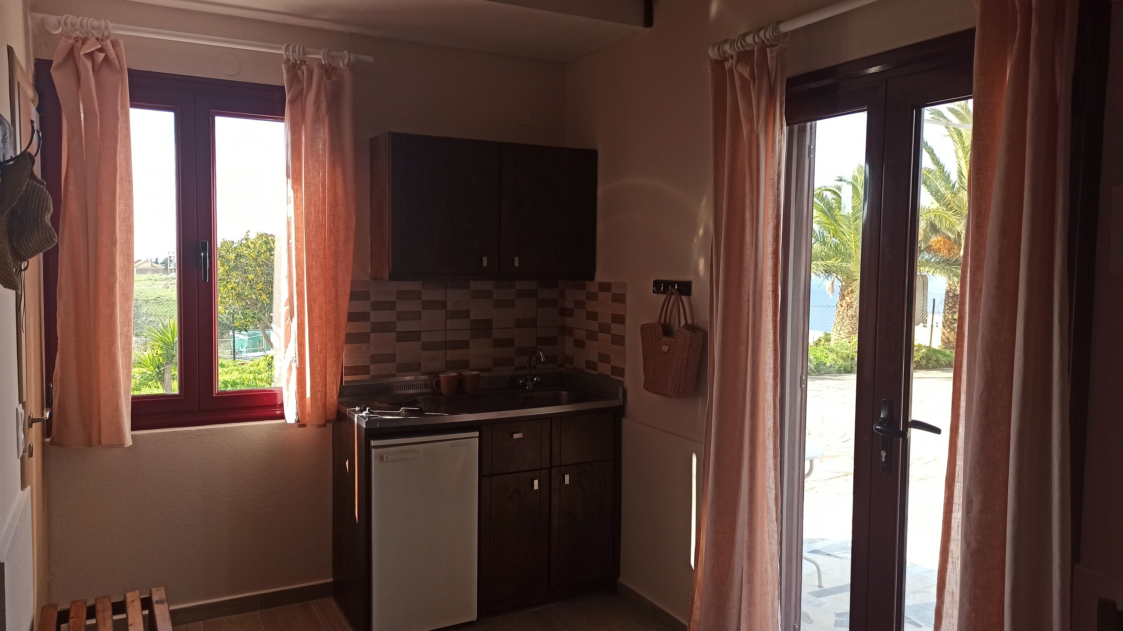 A compact kitchen with a sink, stove, and cabinets, a window with curtains, and a door leading outside.