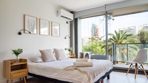 A modern bedroom with a large bed, a nightstand, a chair, and a balcony with a view of buildings and trees.