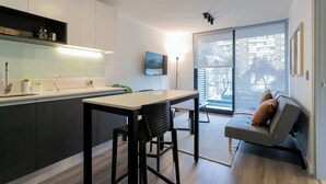 A modern kitchen with a dining area, a sofa with cushions, and a large window offering a view of the city.