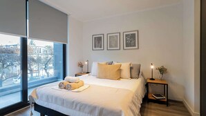 A modern bedroom with a large bed, a nightstand with a lamp, and a view of the city through large windows.
