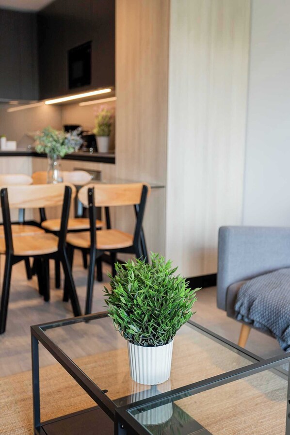 A modern living room with a glass coffee table, a potted plant, a dining area with wooden chairs, and a grey sofa.