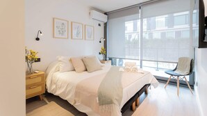 Un dormitorio moderno con una cama grande, una mesita de noche con lámpara, una silla junto a la ventana y cuadros en la pared.