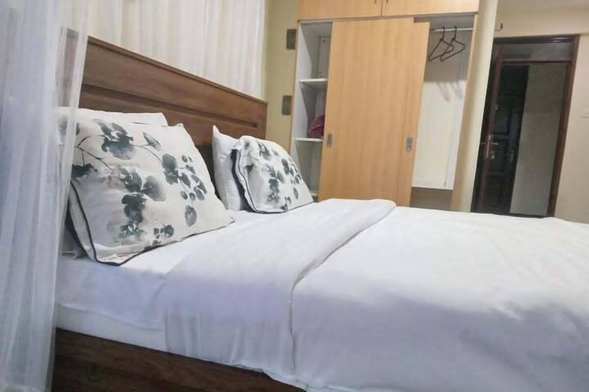 Una cama bien tendida con ropa blanca y almohadas estampadas con motivos florales, cabecera de madera y un armario con espacio para colgar.