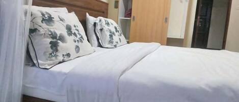 A neatly made bed with white bedding and floral-patterned pillows, a wooden headboard, and a wardrobe with a hanging section.