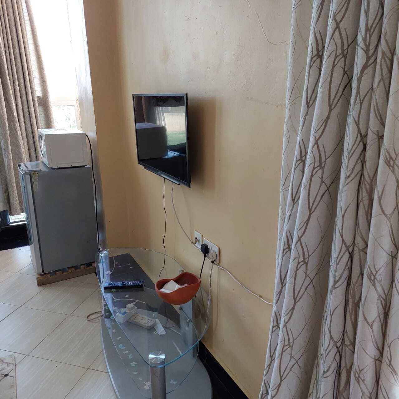 A living room with a flat-screen TV mounted on the wall, a glass coffee table with a remote control, a small refrigerator, and a patterned curtain.