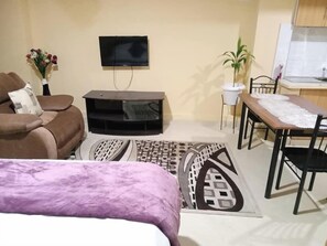 A living room with a sofa, a TV mounted on the wall, a dining table with chairs, and a small kitchen area.