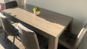 Una mesa rectangular de comedor con acabado en madera clara, acompañada de cuatro sillas tapizadas con tela oscura. Una pequeña planta en maceta se encuentra sobre la mesa.