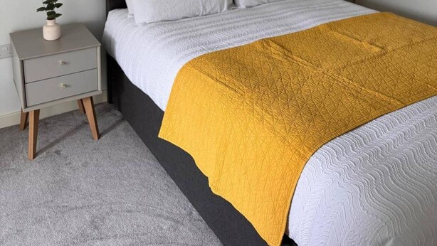 Una cama con colcha blanca y amarilla, una mesita de noche gris con cajones y una pequeña planta en maceta sobre la mesita.