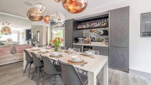 A modern dining area with a long table set for six, a kitchen with a bar area, and a living space with a sofa and a dining table. - Spacious Family Home near Windsor - By Tempstay (Berkshire)