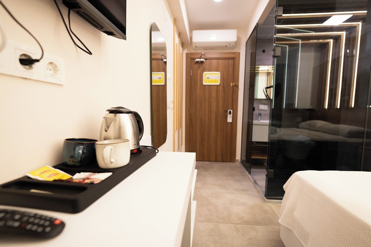 A modern hotel room with a bed, a bedside table with a kettle, a cup, and a remote control. There is a TV mounted on the wall, a door with a mirror, and a glass partition.