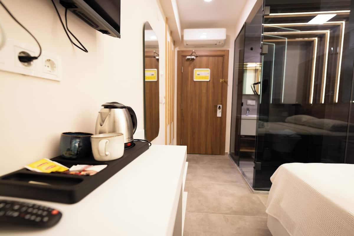A modern hotel room with a bed, a bedside table with a kettle, a cup, and a remote control. There is a TV mounted on the wall, a door with a mirror, and a glass partition.