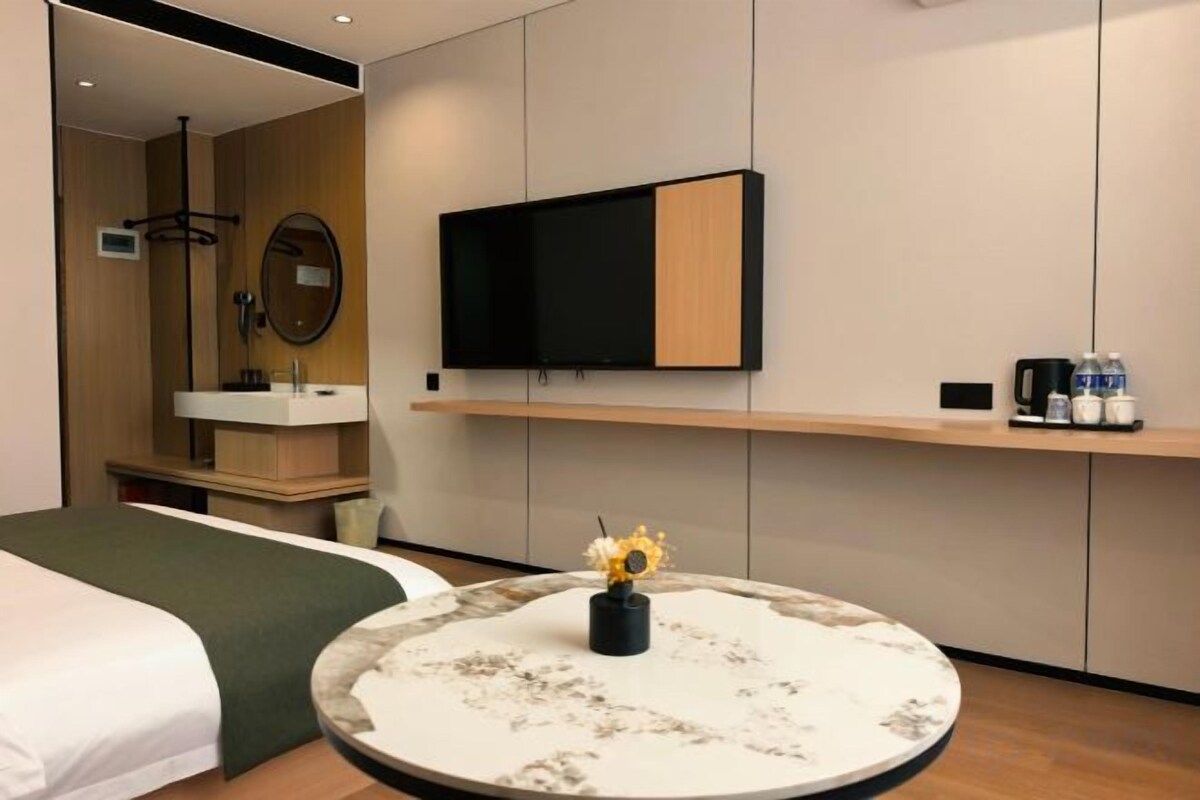 A hotel room with a bed, a round table with a vase, a television mounted on the wall, and a small shelf with items.