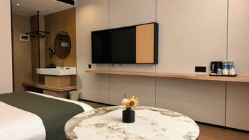 Habitación de hotel con cama, una mesa redonda con un florero, un televisor montado en la pared y un estante con objetos.