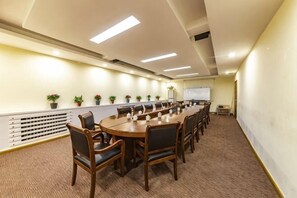 A conference room with a long table, chairs, potted plants, a whiteboard, and a projector screen.