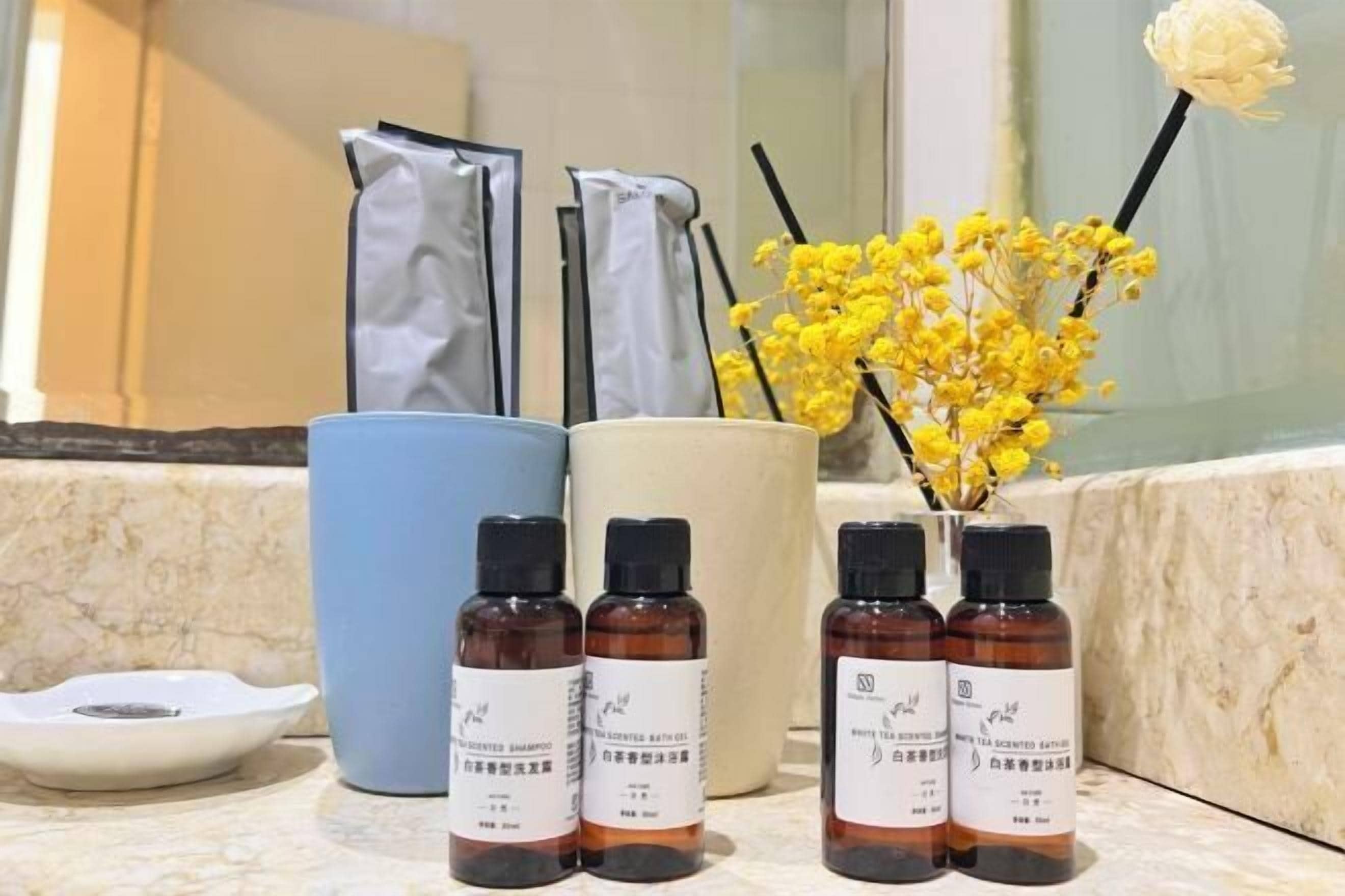 Ein Badezimmer-Waschtisch mit drei braunen Flaschen parfümierten Produkts, einer weißen Schale und einer Vase mit gelben Blumen.