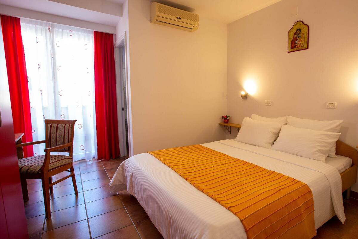 A hotel room with a bed, a chair, a window with red curtains, and a wall-mounted air conditioner.