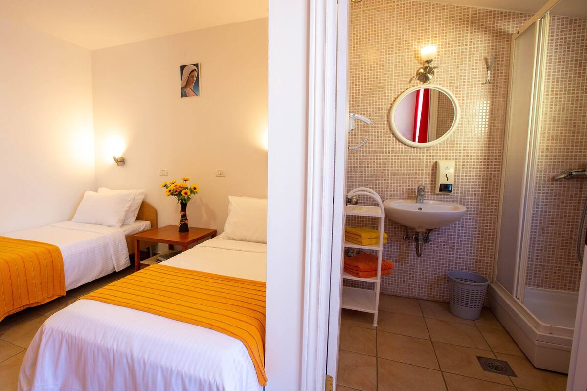 A hotel room with two single beds, a wooden nightstand with a vase of sunflowers, a bathroom with a sink, mirror, and shower.