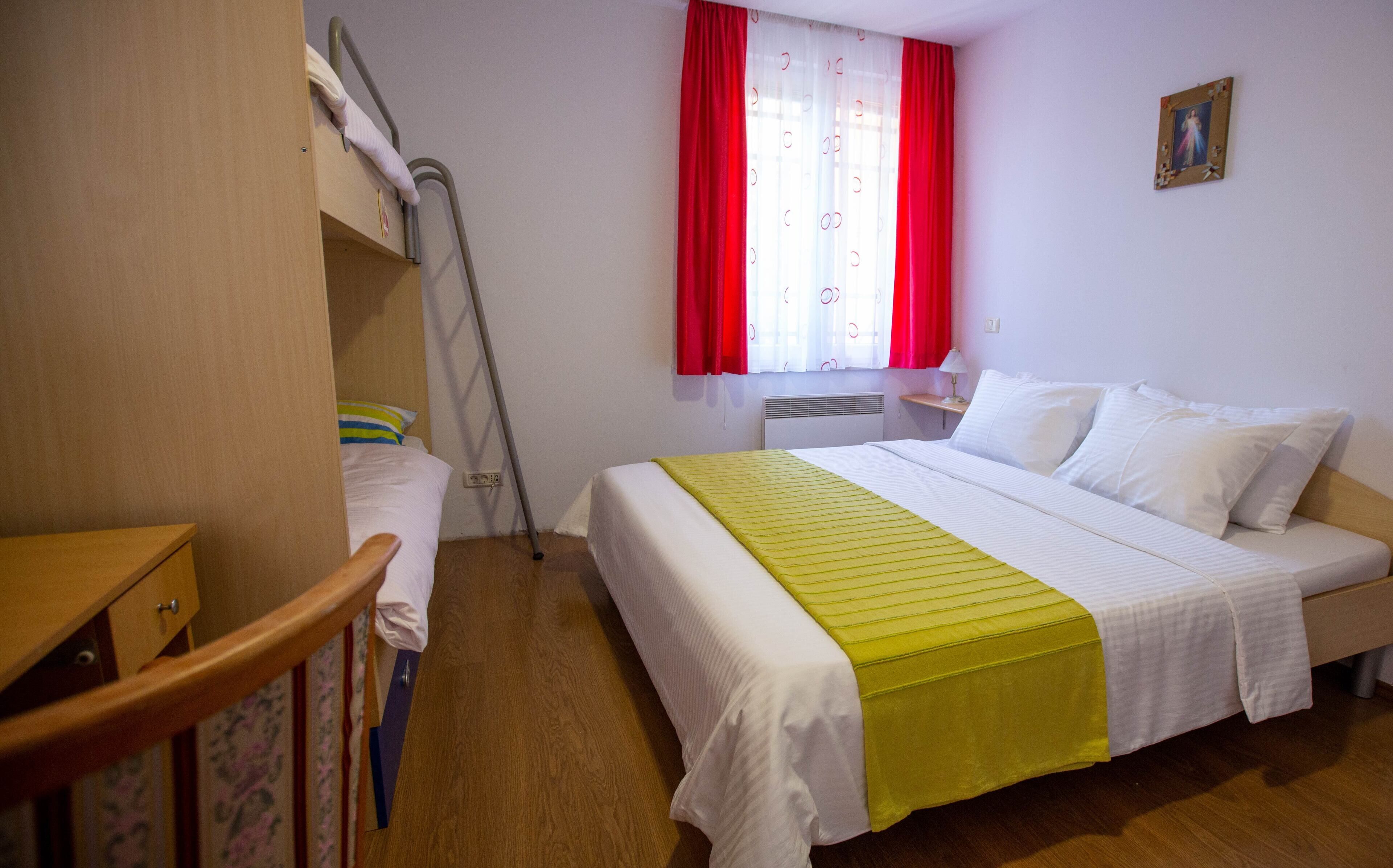 A hotel room with a bunk bed, a single bed, wooden flooring, a bedside table with a lamp, a window with red curtains, and a wall-mounted picture.