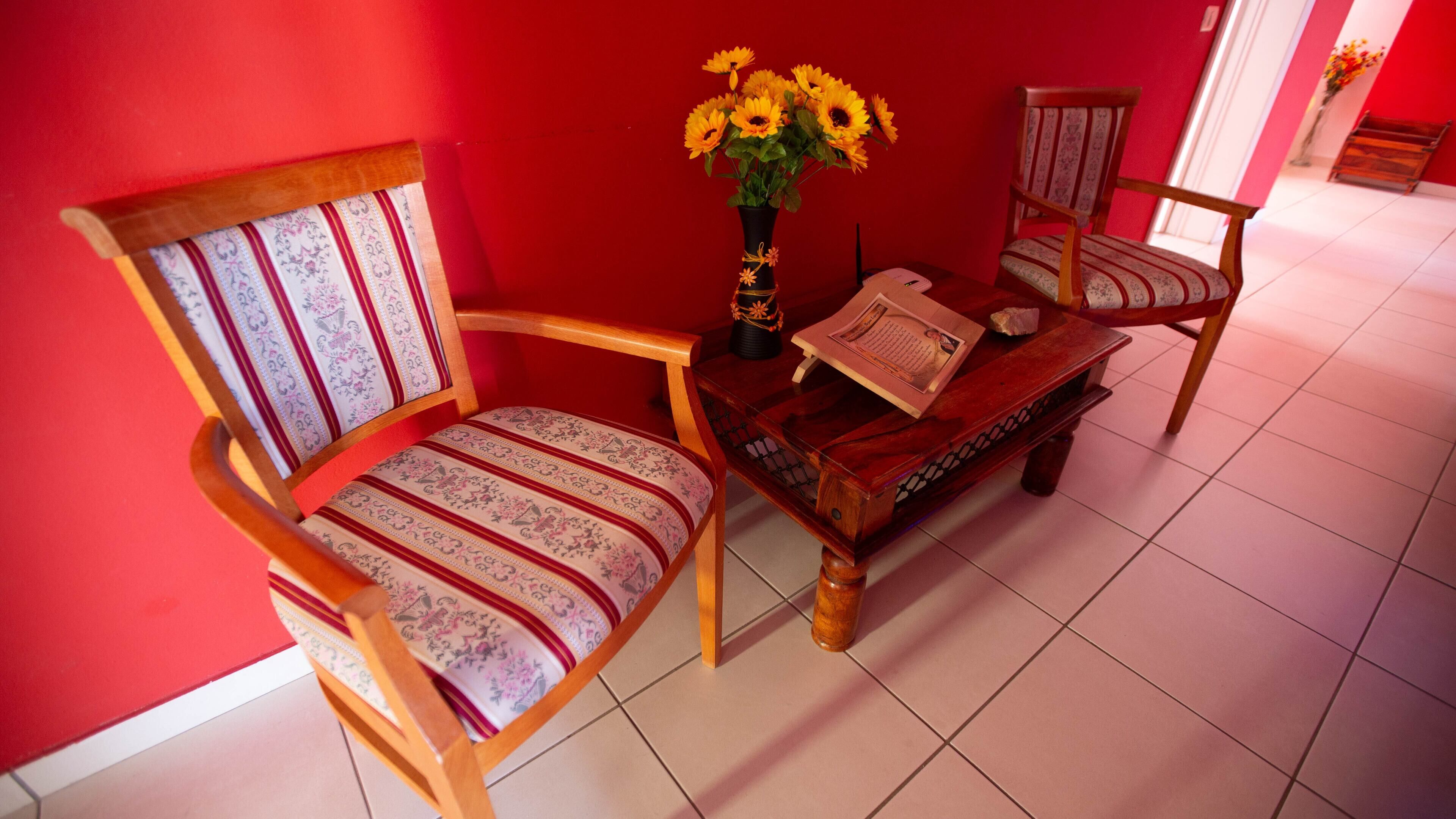 A room with a red wall, a wooden chair with a patterned cushion, a wooden table with a vase of yellow flowers, and a wooden bench.