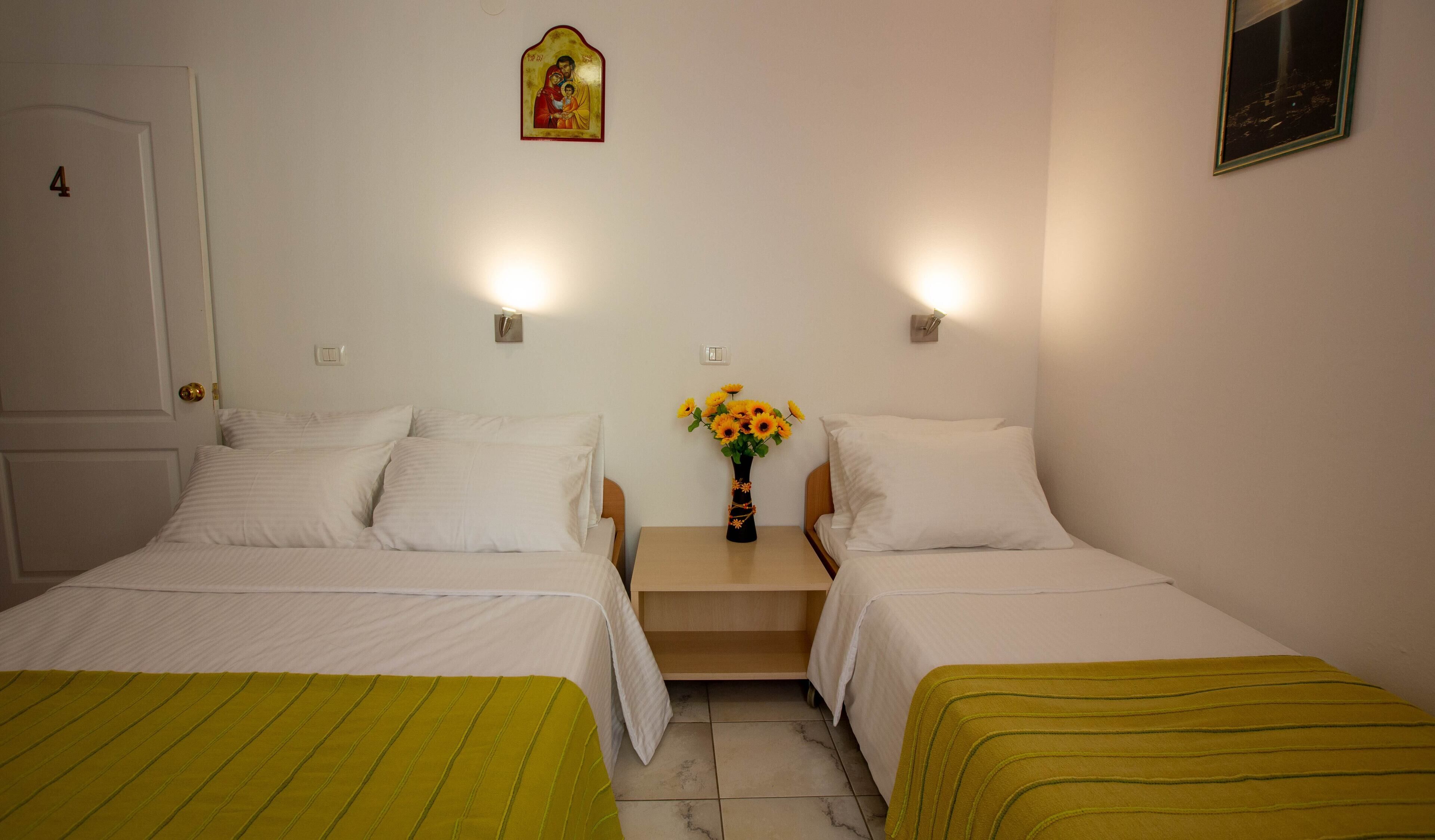 A hotel room with two beds, a nightstand with a vase of flowers, and a religious icon on the wall.