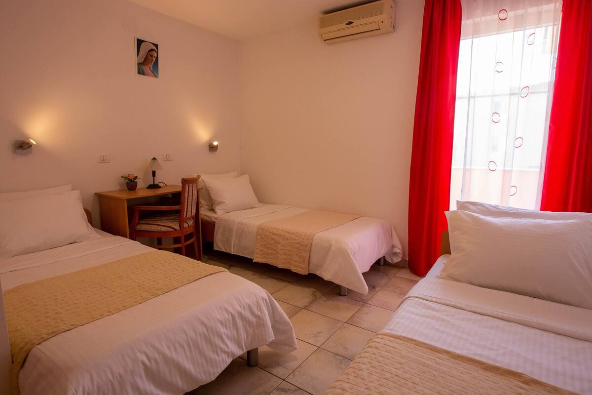 A hotel room with two single beds, a wooden desk, a chair, a small table with a lamp, and a wall-mounted air conditioner.