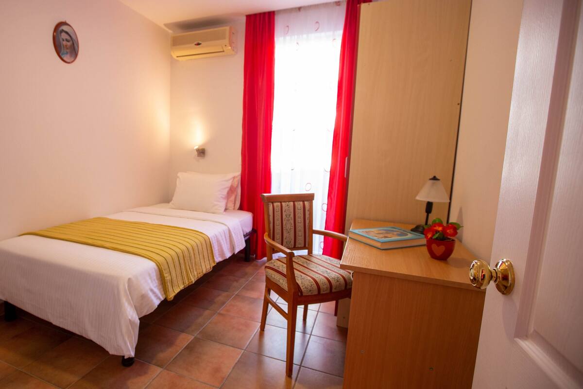 A bedroom with a bed, a chair, a desk, a lamp, and a window with red curtains.