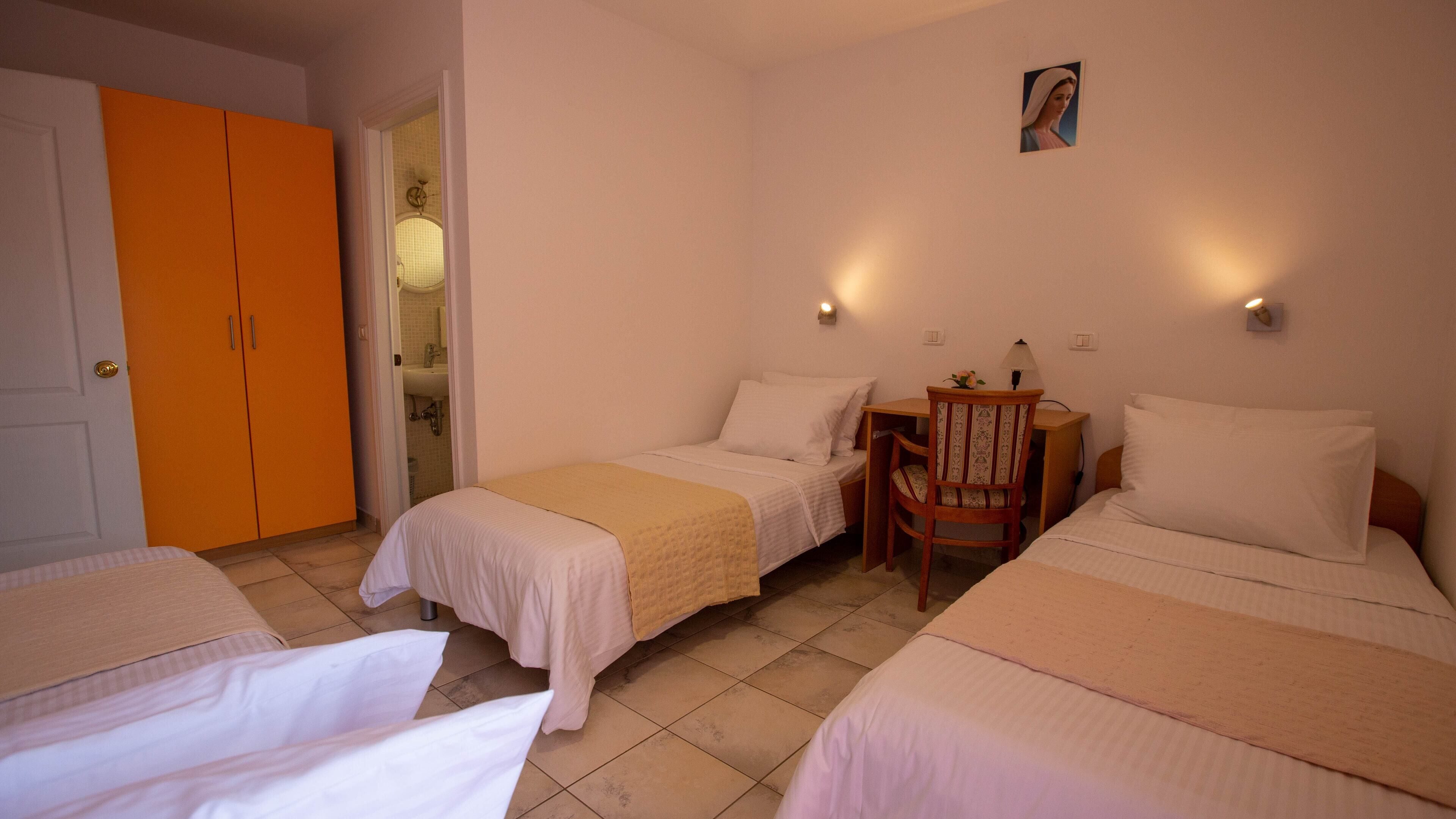 Quarto de hotel com duas camas de solteiro, uma cadeira de madeira, uma mesa pequena e um banheiro visível através de uma porta aberta.