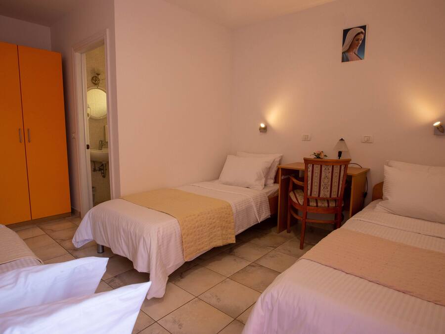 Quarto de hotel com duas camas de solteiro, uma cadeira de madeira, uma mesa pequena e um banheiro visível através de uma porta aberta.