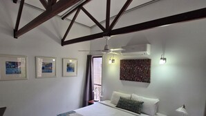A hotel room with a large bed, ceiling fan, air conditioning unit, framed pictures, and a window with curtains.