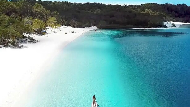 A beach with clear turquoise water and a person paddleboarding.