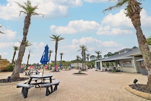 A paved outdoor area with picnic tables, umbrellas, and palm trees.