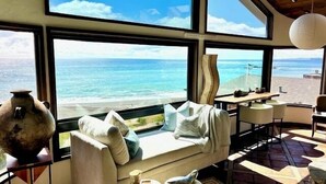 A room with a large window overlooking a beach, a sofa with cushions, a table, and a hanging light fixture.
