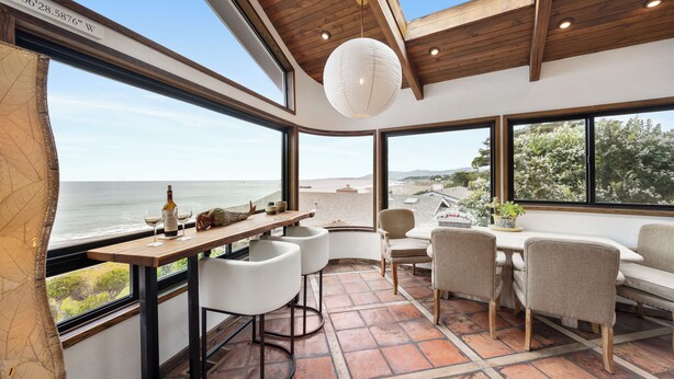 A modern interior with a wooden ceiling, large windows, a dining area with white chairs, and a view of the ocean.
