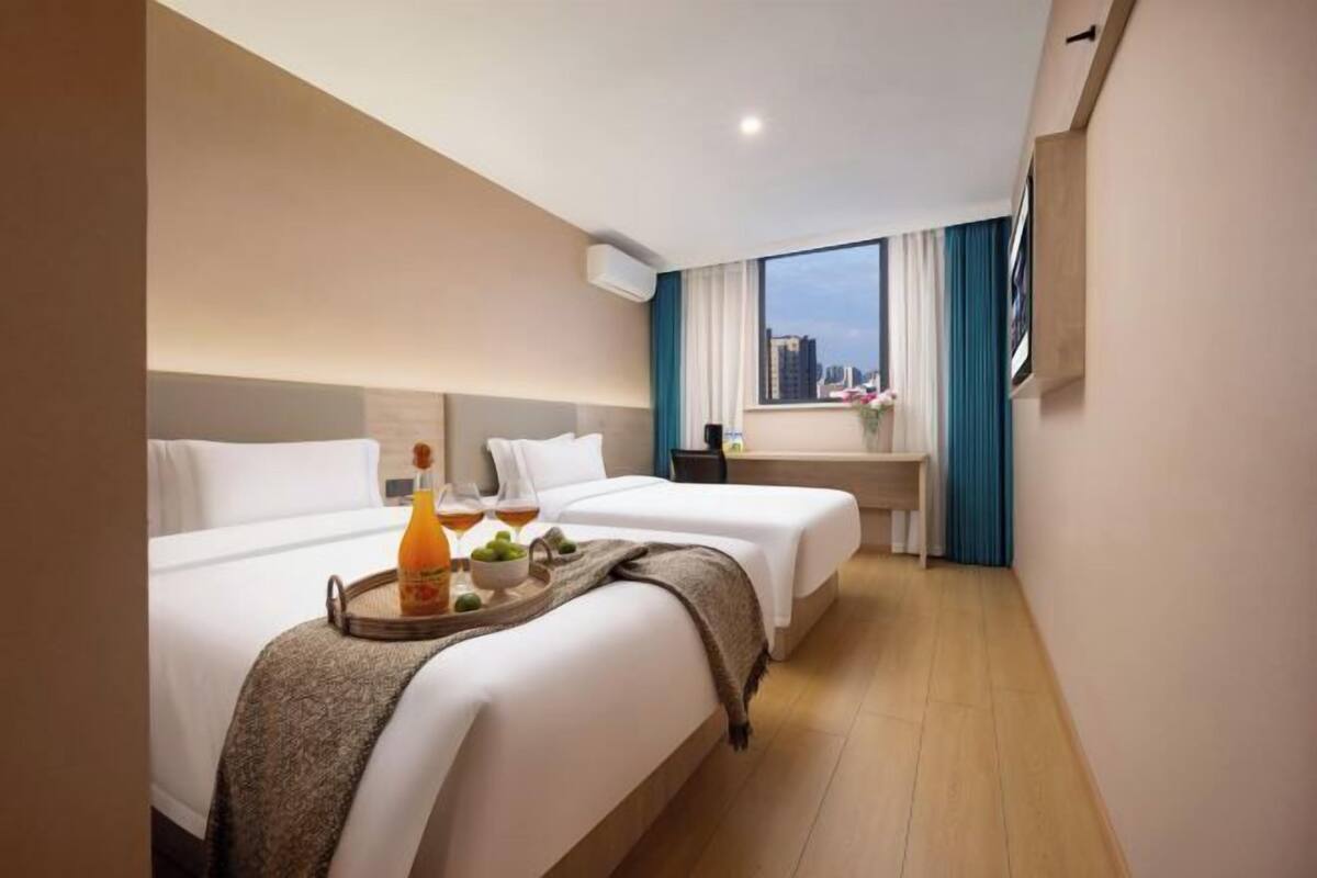 A hotel room with two beds, a small desk, a window with curtains, and a tray with drinks and fruit on the bed.