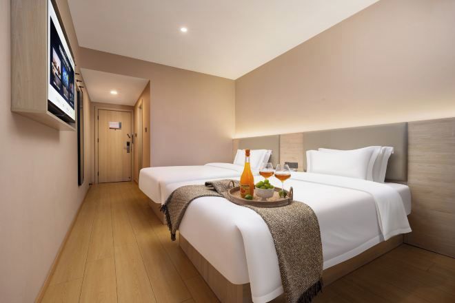 Quarto de hotel com duas camas, piso de madeira, televisão fixada na parede e um recipiente com garrafa e copos sobre a cama.