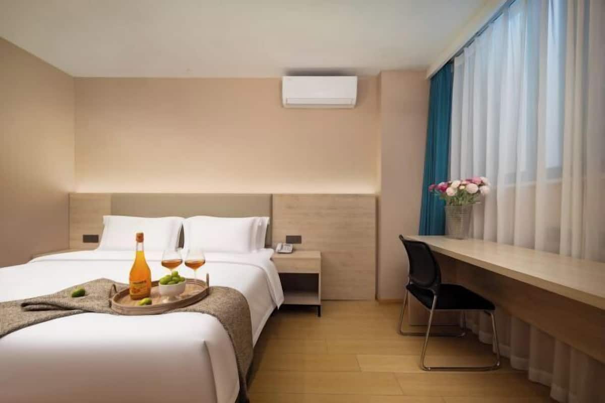 A hotel room with a bed, bedside table, a desk with a chair, a window with curtains, and a wall-mounted air conditioner.