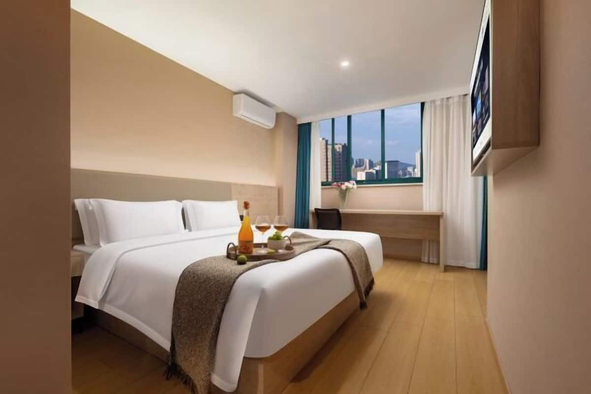A hotel room with a bed, a bedside table, a window with curtains, and a city view.