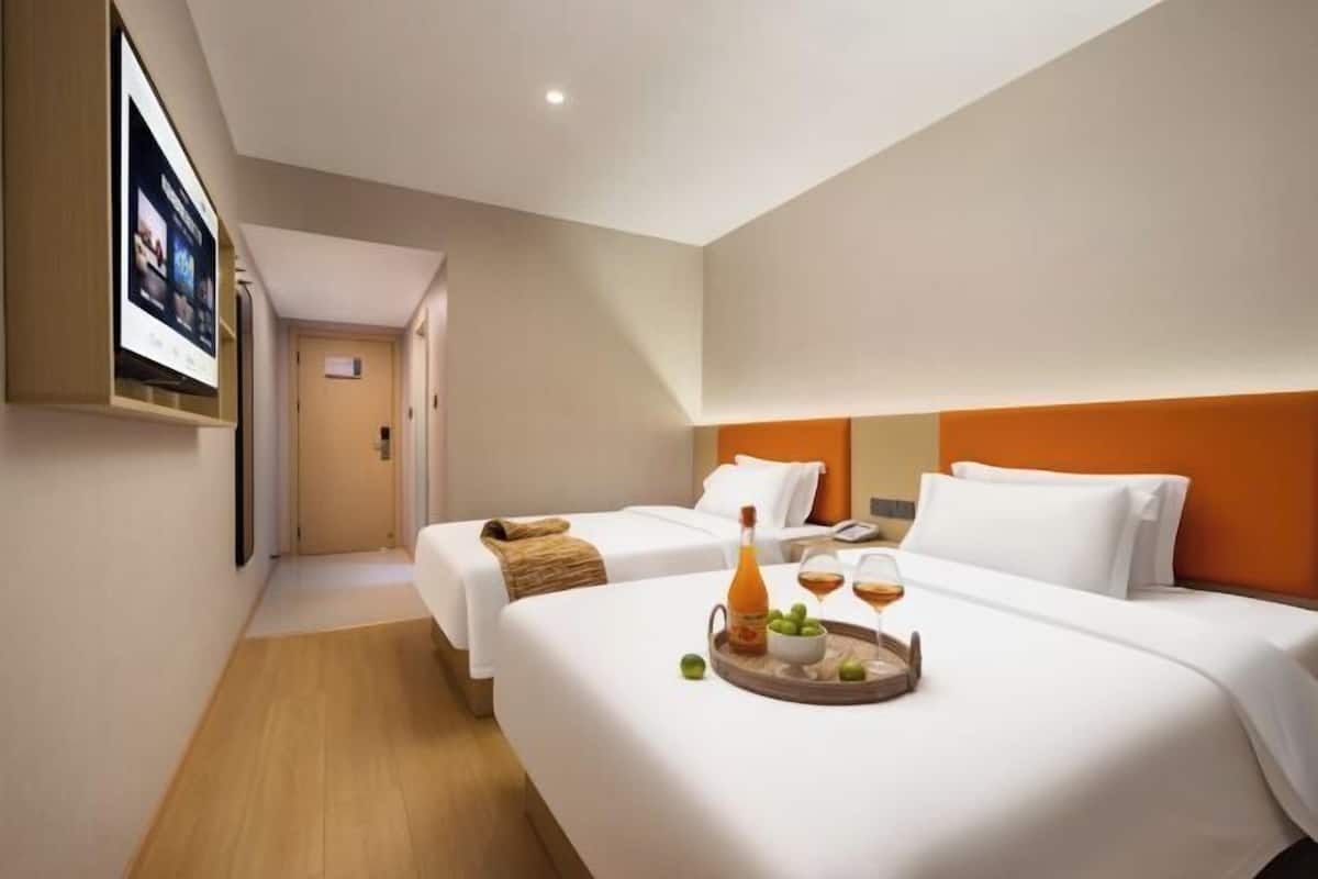 A hotel room with two beds, a wooden floor, a TV mounted on the wall, and a tray with a bottle and glasses on the bed.