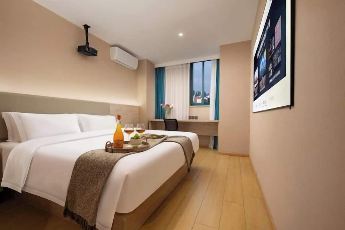 A hotel room with a bed, a desk, a chair, a window with curtains, and a wall-mounted air conditioner.