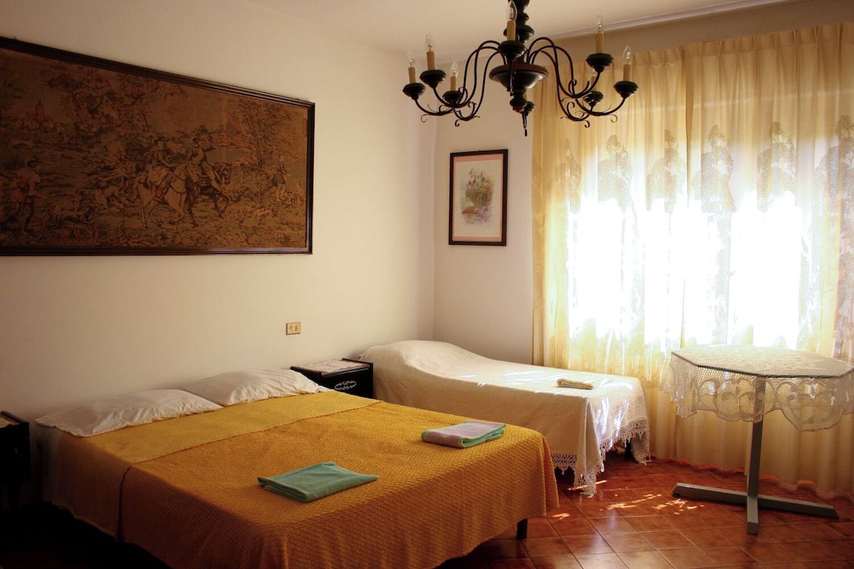 A room with two beds, a table, a chandelier, and a large painting on the wall.
