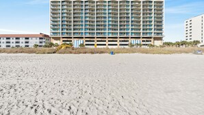 A beachfront hotel with multiple balconies, a sandy beach in the foreground, and a clear sky.