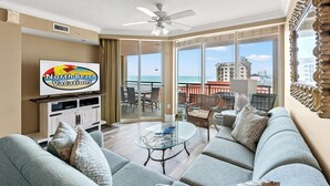 A spacious living room with a large sofa, a glass coffee table, and a television displaying 'North Beach Vacations'. A ceiling fan is visible, and the room has large windows offering a view of the ocean and cityscape.