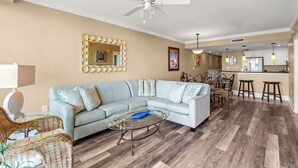 A living room with a light blue sofa, a glass coffee table, wicker chairs, a ceiling fan, and a kitchen area with stools and a bar.