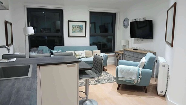 A modern living room with a sofa, a small table, and a TV. There is a kitchen area with a sink and cabinets. - Seaview Loft 20 Portrush (Portrush)